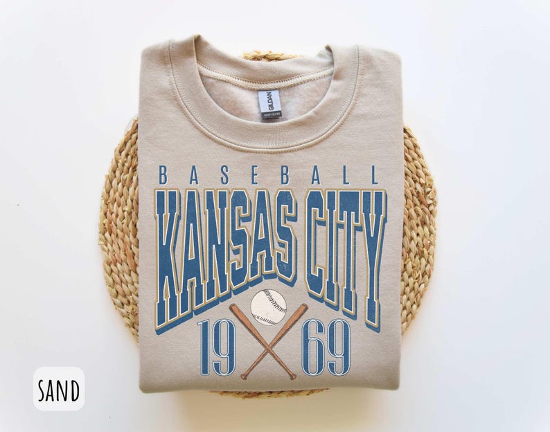 Peut inclure: Un sweat-shirt beige avec le texte "BASEBALL KANSAS CITY 19 69" en lettres bleues et dor&eacute;es. Le texte est entour&eacute; d'un ballon de baseball et de chauves-souris crois&eacute;es.
