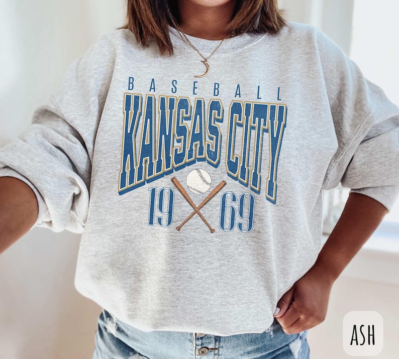 Peut inclure: Sweat-shirt gris avec un motif bleu et or qui dit "Baseball Kansas City 19 69" avec un ballon de baseball et des chauves-souris crois&eacute;es.
