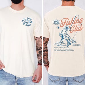May include: Cream-coloured t-shirt with a fishing theme. The front features a blue graphic of a fish and the text "Reel Cool Dad." The back displays a fisherman graphic and the words "Fishing Club Since 2026."