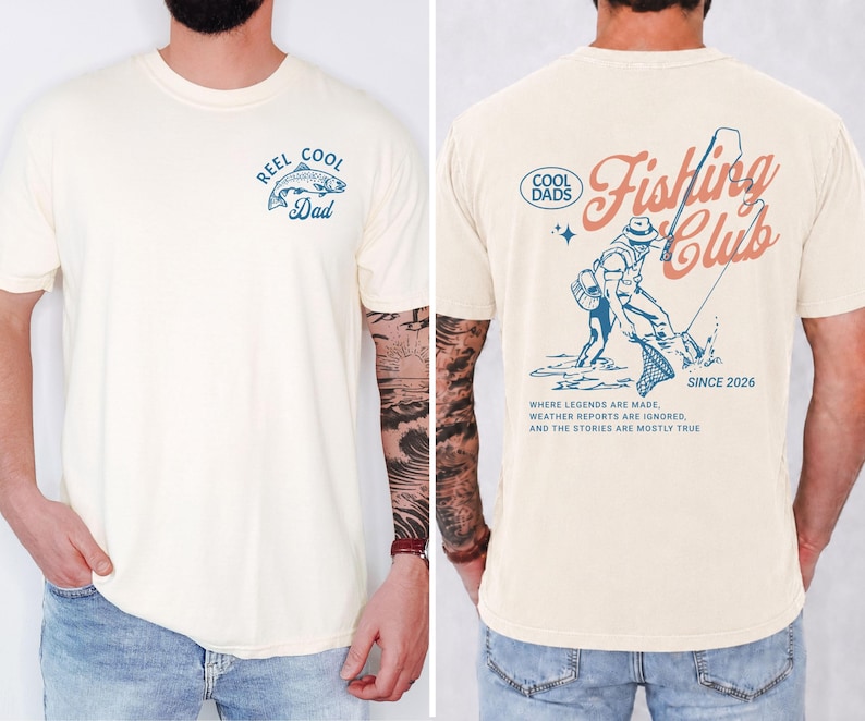 May include: Cream-coloured t-shirt with a fishing theme. The front features a blue graphic of a fish and the text "Reel Cool Dad." The back displays a fisherman graphic and the words "Fishing Club Since 2026."