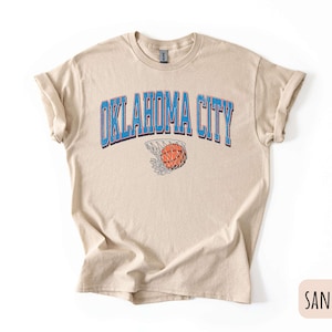 May include: A sand coloured t-shirt with a blue and white graphic of a basketball going through a hoop with the text "OKLAHOMA CITY" above the basketball.