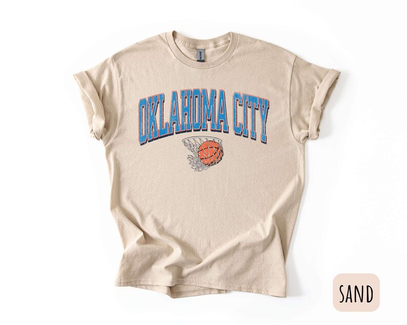May include: A sand coloured t-shirt with a blue and white graphic of a basketball going through a hoop with the text "OKLAHOMA CITY" above the basketball.