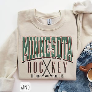 Peut inclure: Un sweat-shirt cr&egrave;me avec le texte "Minnesota Hockey Est 1997" en lettres vertes, rouges et brunes. Le sweat-shirt pr&eacute;sente un graphique de deux b&acirc;tons de hockey crois&eacute;s avec une rondelle.