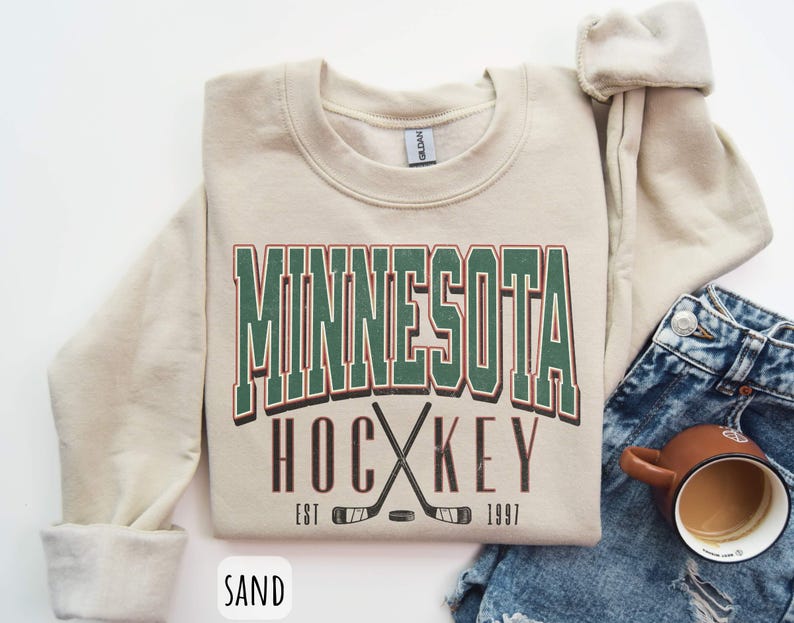 Peut inclure: Un sweat-shirt cr&egrave;me avec le texte "Minnesota Hockey Est 1997" en lettres vertes, rouges et brunes. Le sweat-shirt pr&eacute;sente un graphique de deux b&acirc;tons de hockey crois&eacute;s avec une rondelle.
