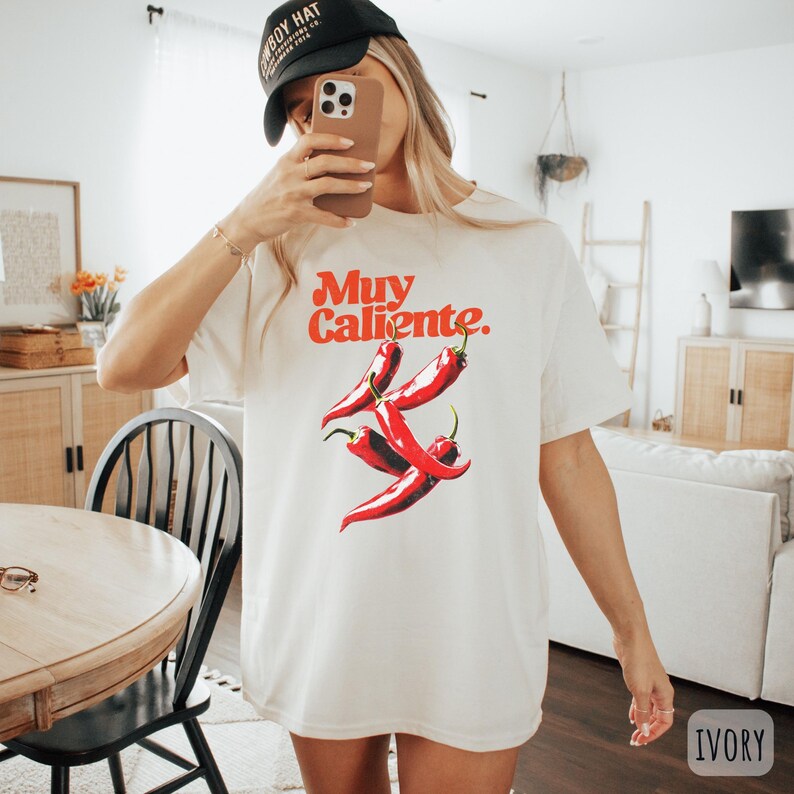 May include: Ivory-colored t-shirt with the text "Muy Caliente" in red, along with a graphic of three red chili peppers. The shirt is a casual style, suitable for everyday wear.