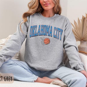 May include: A grey sweatshirt with the text "OKLAHOMA CITY" in blue and a basketball in a hoop graphic. The sweatshirt is being worn by a person sitting on a sofa.