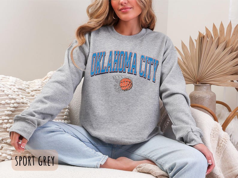 May include: A grey sweatshirt with the text "OKLAHOMA CITY" in blue and a basketball in a hoop graphic. The sweatshirt is being worn by a person sitting on a sofa.