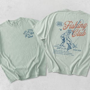 May include: Two light green t-shirts. One features a fish graphic and the text "REEL COOL Dad". The other displays a fisherman graphic and the words "COOL DADS Fishing Club SINCE 2026". The text "WHERE LEGENDS ARE MADE, WEATHER REPORTS ARE IGNORED, AND THE STORIES ARE MOSTLY TRUE" is also printed on the shirt.