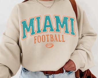 May include: A beige crewneck sweatshirt featuring the text MIAMI in distressed teal letters with orange outlines, followed by the word FOOTBALL in orange, and a small orange football graphic centered below the text on the front of the garment.