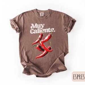 May include: Espresso-colored t-shirt with the words "Muy Caliente" in white script above a graphic of four red chili peppers. The shirt has rolled-up sleeves and a crew neck.