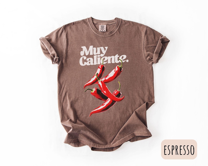 May include: Espresso-colored t-shirt with the words "Muy Caliente" in white script above a graphic of four red chili peppers. The shirt has rolled-up sleeves and a crew neck.
