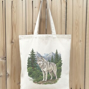 Op de afbeelding: Cr&egrave;mekleurige draagtas met een gedetailleerde illustratie van een grijze wolf in een boslandschap. Het ontwerp bevat groenblijvende bomen, bergen en groen, waardoor een accessoire met een natuurthema ontstaat.