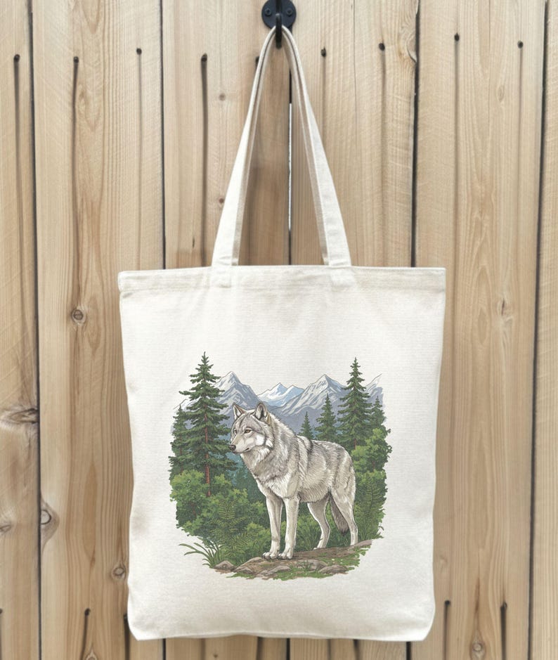Op de afbeelding: Cr&egrave;mekleurige draagtas met een gedetailleerde illustratie van een grijze wolf in een boslandschap. Het ontwerp bevat groenblijvende bomen, bergen en groen, waardoor een accessoire met een natuurthema ontstaat.