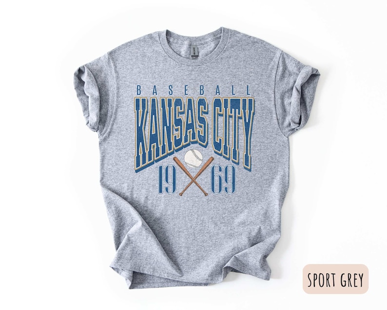 Peut inclure: Un t-shirt gris avec un design graphique de style vintage repr&eacute;sentant le texte "Baseball Kansas City 1969" avec un ballon de baseball et des chauves-souris crois&eacute;es.