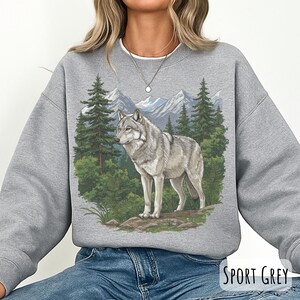 Op de afbeelding: Sportgrijs sweatshirt met een wolf illustratie in een boslandschap. Het ontwerp bevat een wolf, groenblijvende bomen en bergen. De sweatshirt heeft een ronde hals. De tekst "Sport Grey" is zichtbaar.