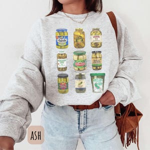 Op de afbeelding: Lichtgrijze sweatshirt met een afbeelding van verschillende augurkenpotten. De potten hebben verschillende vormen en maten, met labels zoals Vlasic en Claussen. De sweatshirt heeft lange mouwen en een ronde hals.