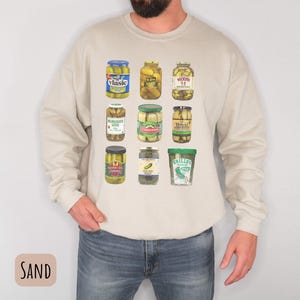 Op de afbeelding: Zandkleurig sweatshirt met lange mouwen met een raster van negen augurkenpotten met verschillende etiketten. De potten zijn gevuld met augurken en hebben verschillend gekleurde deksels. Het sweatshirt is gemaakt van een zacht materiaal.