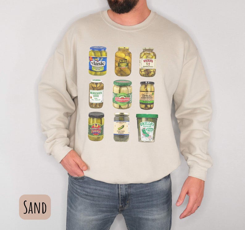 Op de afbeelding: Zandkleurig sweatshirt met lange mouwen met een raster van negen augurkenpotten met verschillende etiketten. De potten zijn gevuld met augurken en hebben verschillend gekleurde deksels. Het sweatshirt is gemaakt van een zacht materiaal.