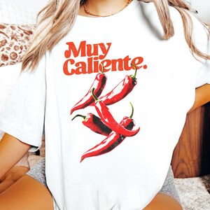 May include: White t-shirt with a red graphic of chili peppers and the words "Muy Caliente." The design features four red chili peppers with green stems. The text is in a bold, retro font.