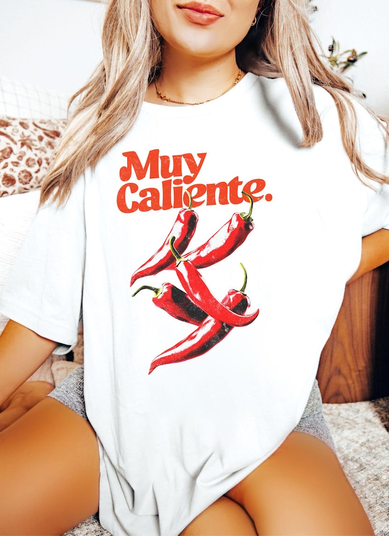 May include: White t-shirt with a red graphic of chili peppers and the words "Muy Caliente." The design features four red chili peppers with green stems. The text is in a bold, retro font.