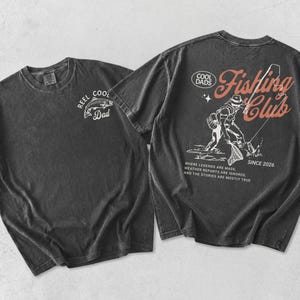 May include: Two charcoal grey t-shirts. One has a white graphic of a fish and the text "REEL COOL Dad". The other features a fishing-themed design with the words "Fishing Club" and "Since 2026". The text "Where legends are made, weather reports are ignored, and the stories are mostly true" is also included.