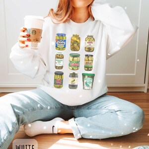 Op de afbeelding: Witte sweatshirt met een kleurrijke afbeelding van verschillende augurkenpotten. De potten zijn gevuld met augurken en hebben verschillende merklabels. Het woord "WHITE" staat onderaan de afbeelding.