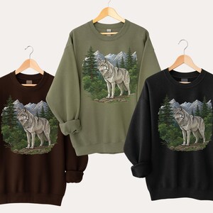 Op de afbeelding: Drie sweatshirts in bruin, olijfgroen en zwart, elk met een wolf illustratie. De wolf staat in een boslandschap met bergen op de achtergrond. De sweatshirts hangen aan houten hangers.