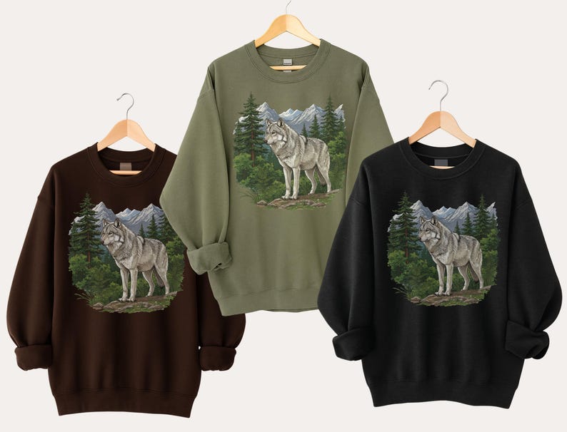 Op de afbeelding: Drie sweatshirts in bruin, olijfgroen en zwart, elk met een wolf illustratie. De wolf staat in een boslandschap met bergen op de achtergrond. De sweatshirts hangen aan houten hangers.