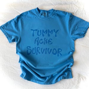 May include: A blue t-shirt with the words "TUMMY ACHE SURVIVOR" printed in a darker blue. The shirt is made of a soft material and has a crew neck. The shirt is laying on a white surface.