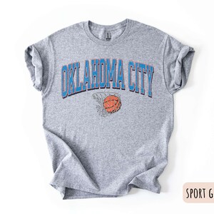 May include: A grey t-shirt with the text "OKLAHOMA CITY" in blue letters and a basketball going through a hoop in the centre of the text. The shirt is a sport grey colour.