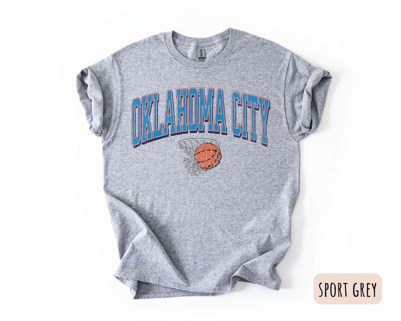 May include: A grey t-shirt with the text "OKLAHOMA CITY" in blue letters and a basketball going through a hoop in the centre of the text. The shirt is a sport grey colour.