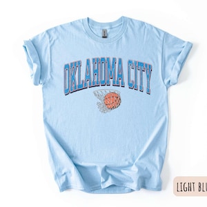 May include: Light blue t-shirt with the text "OKLAHOMA CITY" in blue and red lettering. A basketball is shown going through a hoop.
