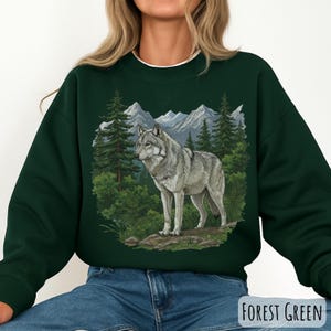 Op de afbeelding: Donkergroene sweatshirt met een illustratie van een grijze wolf in een boslandschap met bergen op de achtergrond. De tekst "FOREST GREEN" staat rechtsonder. De sweatshirt heeft een witte kraag.