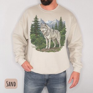 Op de afbeelding: Zandkleurige sweatshirt met een gedetailleerde illustratie van een grijze wolf in een boslandschap met bergen. De wolf is het middelpunt, tegen een achtergrond van groene bomen en een blauwe lucht. De sweatshirt heeft lange mouwen en een ronde hals. Het woord "SAND" staat in een kader.