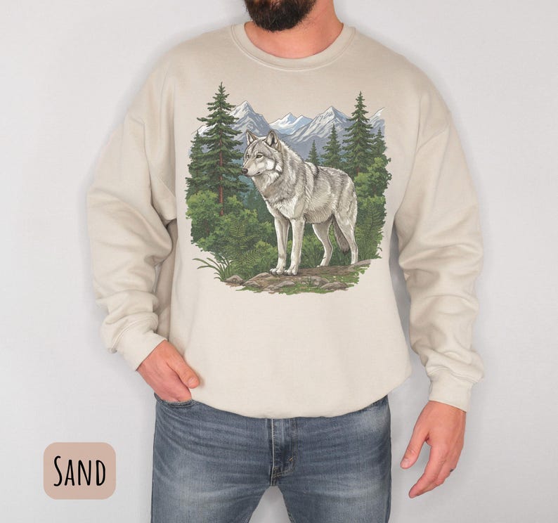 Op de afbeelding: Zandkleurige sweatshirt met een gedetailleerde illustratie van een grijze wolf in een boslandschap met bergen. De wolf is het middelpunt, tegen een achtergrond van groene bomen en een blauwe lucht. De sweatshirt heeft lange mouwen en een ronde hals. Het woord "SAND" staat in een kader.