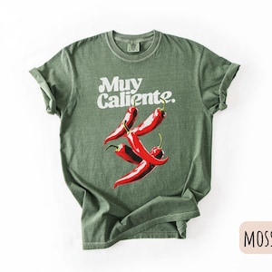 May include: Moss green t-shirt with the words "Muy Caliente" in white script above a graphic of four red chili peppers. The shirt has short sleeves and a crew neck.