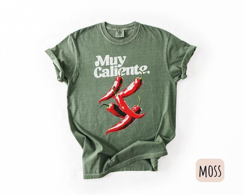 May include: Moss green t-shirt with the words "Muy Caliente" in white script above a graphic of four red chili peppers. The shirt has short sleeves and a crew neck.
