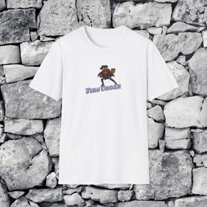 Może przedstawiać: Biały t-shirt z krótkim rękawem z grafiką przedstawiającą osobę w czerwonej pelerynie i niebieskich dżinsach, trzymającą laptopa. Pod postacią znajduje się napis VIBE CODER wydrukowany niebieską, postarzaną czcionką blokową na środku klatki.
