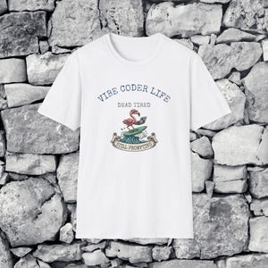 Può includere: Una t-shirt bianca a maniche corte con la grafica di un fenicottero rosa su una tavola da surf che usa un portatile. Il testo recita VIBE CODER LIFE, DEAD TIRED e STILL PROMPTING su un banner sotto l'onda in blu, rosa e verde.
