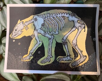 May include: A sticker featuring a black and white illustration of a bear skeleton with a yellow and blue gradient. The bear is outlined in black and has stars on its bones. The background is black with white dots.