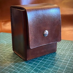 May include: A rectangular brown leather case with a vertical orientation, featuring a fold-over flap secured by a single silver-toned metal snap button. The leather has a smooth, polished finish with visible stitching along the side edges.