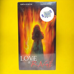 Peut inclure: Pochette de cassette VHS avec un fond rouge et orange représentant une femme en robe blanche. Le titre du film est "LOVE & RAGE" avec les acteurs Greta Scacchi et Daniel Craig mentionnés au-dessus du titre.
