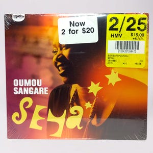 Peut inclure: Un CD de l'album "Seya" d'Oumou Sangare est présenté. La couverture de l'album présente un fond chaud orange et jaune avec un portrait de femme et des graphiques d'étoiles jaunes. Le titre de l'album et le nom de l'artiste sont en grandes lettres grasses.