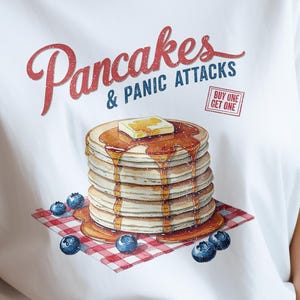 Peut inclure: T-shirt blanc avec l'inscription "Pancakes & Panic Attacks" en rouge et bleu. Une pile de crêpes avec du beurre et du sirop est posée sur une serviette à carreaux rouges et blancs, entourée de myrtilles. Texte "Buy One Get One" dans un encadré rouge.