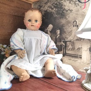 Puede incluir: Una muñeca bebé vintage con rostro pintado, vestida con un vestido blanco con bordados florales azules y rosas. La muñeca está sentada sobre una superficie de madera, con un fondo decorativo con una impresión histórica.