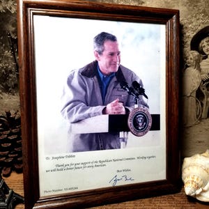 Peut inclure: Une photographie encadrée d'un homme s'exprimant à un podium. L'homme porte une veste marron et une chemise bleue. La photo est signée "Meilleurs vœux, George W. Bush" et comprend le texte "Merci pour votre soutien au Comité national républicain. En travaillant ensemble, nous construirons un avenir meilleur pour tous les Américains."