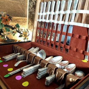 May include: A vintage silverware set in a wooden case with a red velvet interior. The set includes knives, forks, and spoons, neatly arranged. A decorative floral sculpture is visible in the background.