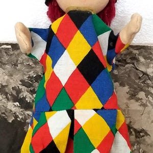 May include: A colorful hand puppet with a red and black checkered pattern. The puppet has a red wig, a black eye mask, and a smiling face.