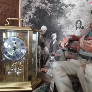 May include: A gold-toned, rectangular clock with a glass front and a light blue clock face. The clock has a handle on top and a pendulum with silver-colored weights. A figurine of an elderly man in a hat is in the background.