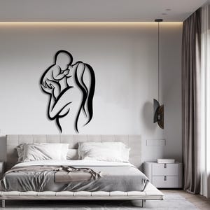 May include: Black silhouette wall art depicting a mother holding a baby. The art is mounted on a white wall in a bedroom setting with a bed, nightstand, and curtains.
